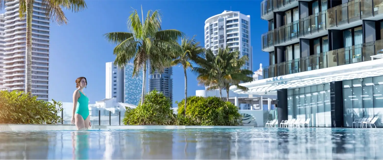 Woman in turquoise swimsuit enjoying luxury poolside with city skyline and palm trees.