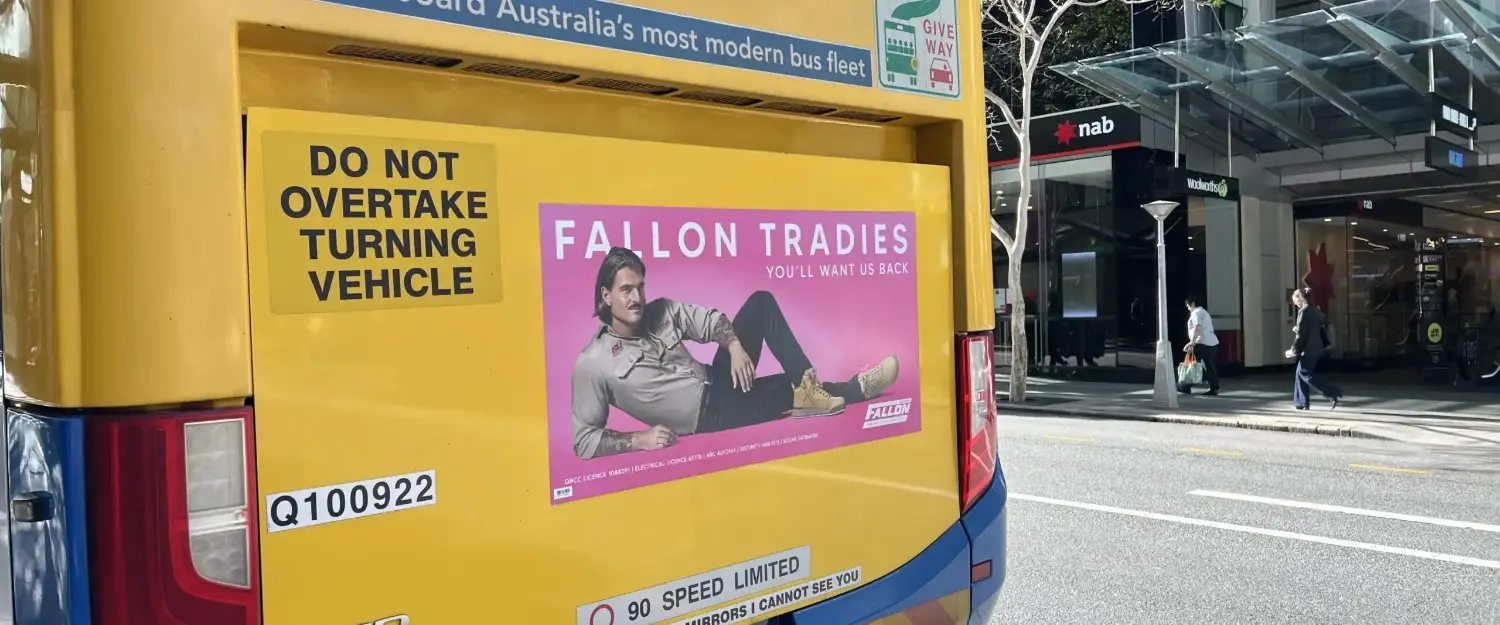 Bright yellow bus featuring Fallon Tradies advertisement in urban setting with pedestrians.